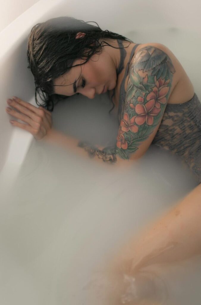 Moody milk bath boudoir session in Las Vegas featuring a woman with floral sleeve tattoos resting against the tub edge.