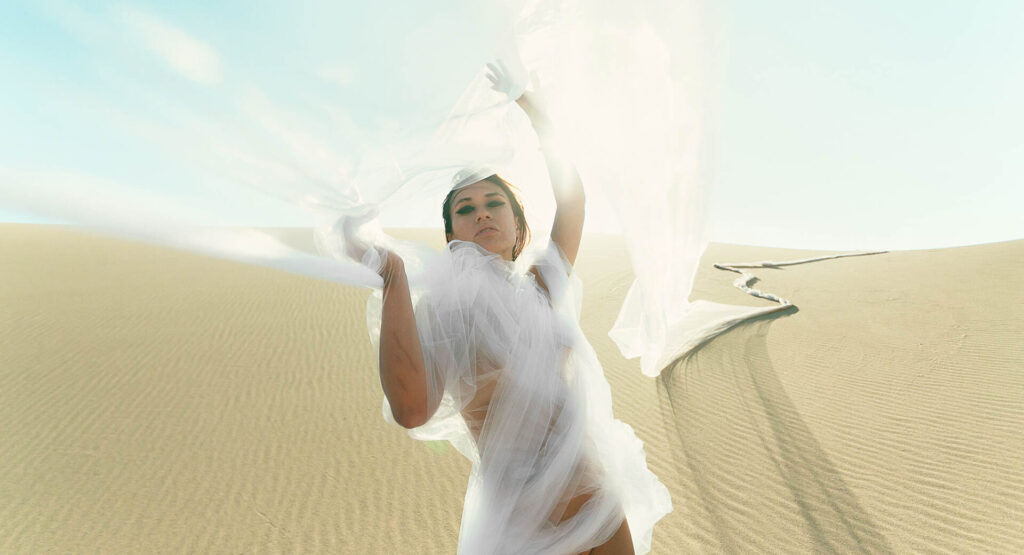 Ethereal desert boudoir session in Las Vegas sand dunes featuring a model wrapped in flowing white tulle fabric.